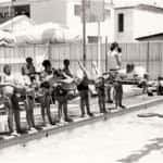 Children learn to swim with the help of instructors at a poolside, using kickboards and wearing swim caps.
