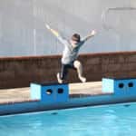 A boy jumps over blue blocks by a swimming pool, with water cascading down a wall in the background.