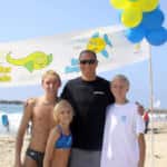A man and two children posing together at a beach under a banner promoting safety for kids, with colorful balloons in the background.