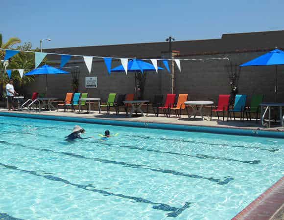 Outdoor swimming pool with children swimming, surrounded by colorful chairs and umbrellas under a clear sky.