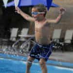 A young boy in swim goggles and blue shorts joyfully jumping near a swimming pool, splashing water around.