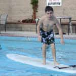 A boy with down syndrome practices surfing on a board in a swimming pool, with other swimmers and a "lifeguard on duty" sign visible in the background.