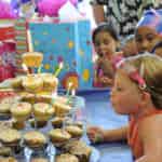 A girl in an orange dress blowing out a candle on cupcakes at a children's birthday party, surrounded by other children and gifts.
