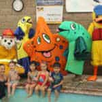 Children and adults posing with colorful sea creature and firefighter mascots by a swimming pool.