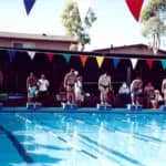 Swimmers preparing to dive into an outdoor pool at a swim meet, with audience onlookers and colorful flags hung above.
