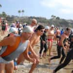 Group of triathletes running into the sea at the start of a race, wearing swim caps and wetsuits, with a rocky beach and cliff in the background.