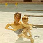 A man and a young child smiling in a swimming pool, sunny day.