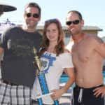Two men and a young woman holding a trophy, smiling; one man is shirtless and the other is in a casual t-shirt, outdoors.