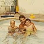 A man with two young boys smiling in a small pool, with a yellow rubber duck floating nearby.