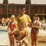 Three children with trophies standing by a swimming pool, accompanied by an adult male. spectators sit in the background.