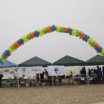 Colorful balloon arch over informational booths on a sandy beach on an overcast day.