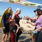 Two adults and a child in wetsuits interact with a woman on a sunny beach, with event banners visible in the background.