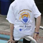 Person standing by a pool wearing a white t-shirt with logos and text "swim for life" and "world record holder" for a swimming event.