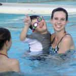 A young boy wearing swim goggles and a float vest points upwards while held by a smiling woman in a swimming pool.