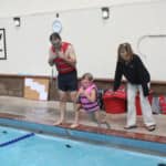 A child wearing a life jacket prepares to enter a swimming pool with the assistance of two adults, one of whom is wearing a life jacket.