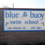 Sign for blue buoy swim school on a brick wall, featuring a blue background, dolphin logo, and the address "1702 nisson rd. est. 1956.
