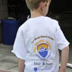 A young boy wearing a white t-shirt that says "the world's largest swimming lesson" with logo, standing by a pool.