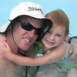 A man wearing a white hat and sunglasses hugging a young girl with red hair in a swimming pool.