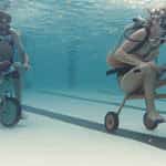Two scuba divers riding underwater bicycles in a clear pool.