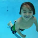 A young boy with a joyful expression swimming underwater in a clear blue pool.