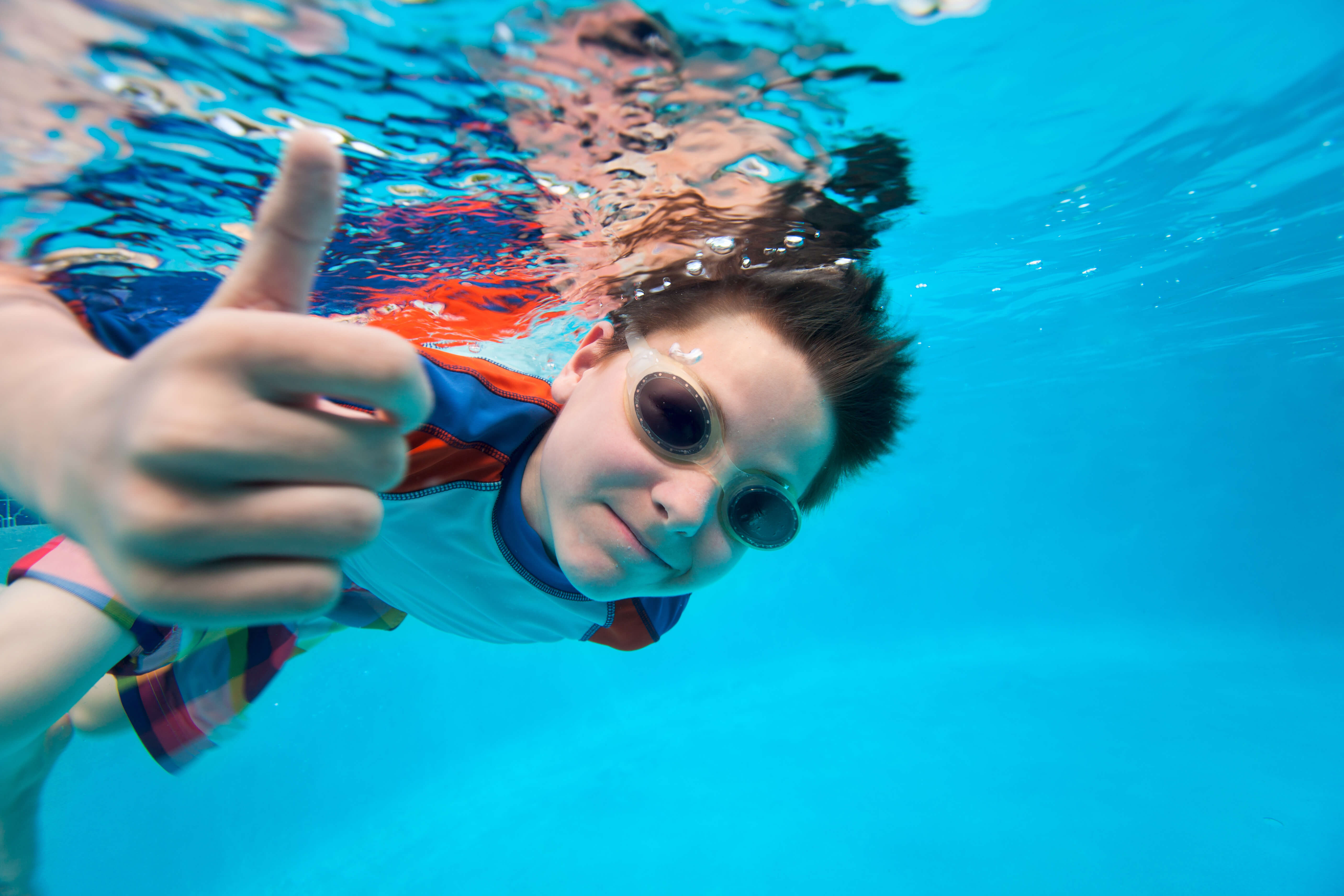 Young boy with goggles giving a thumbs-up underwater in a Huntington Beach swimming pool.