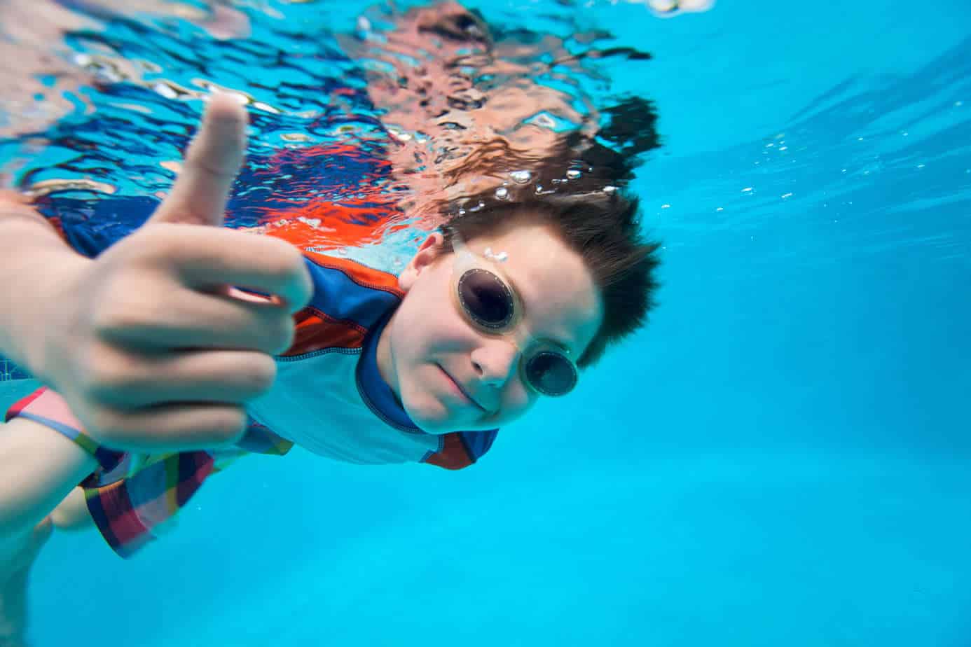 A young boy wearing goggles and a life jacket gives a thumbs up while submerged in the clear blue water of Newport Beach.