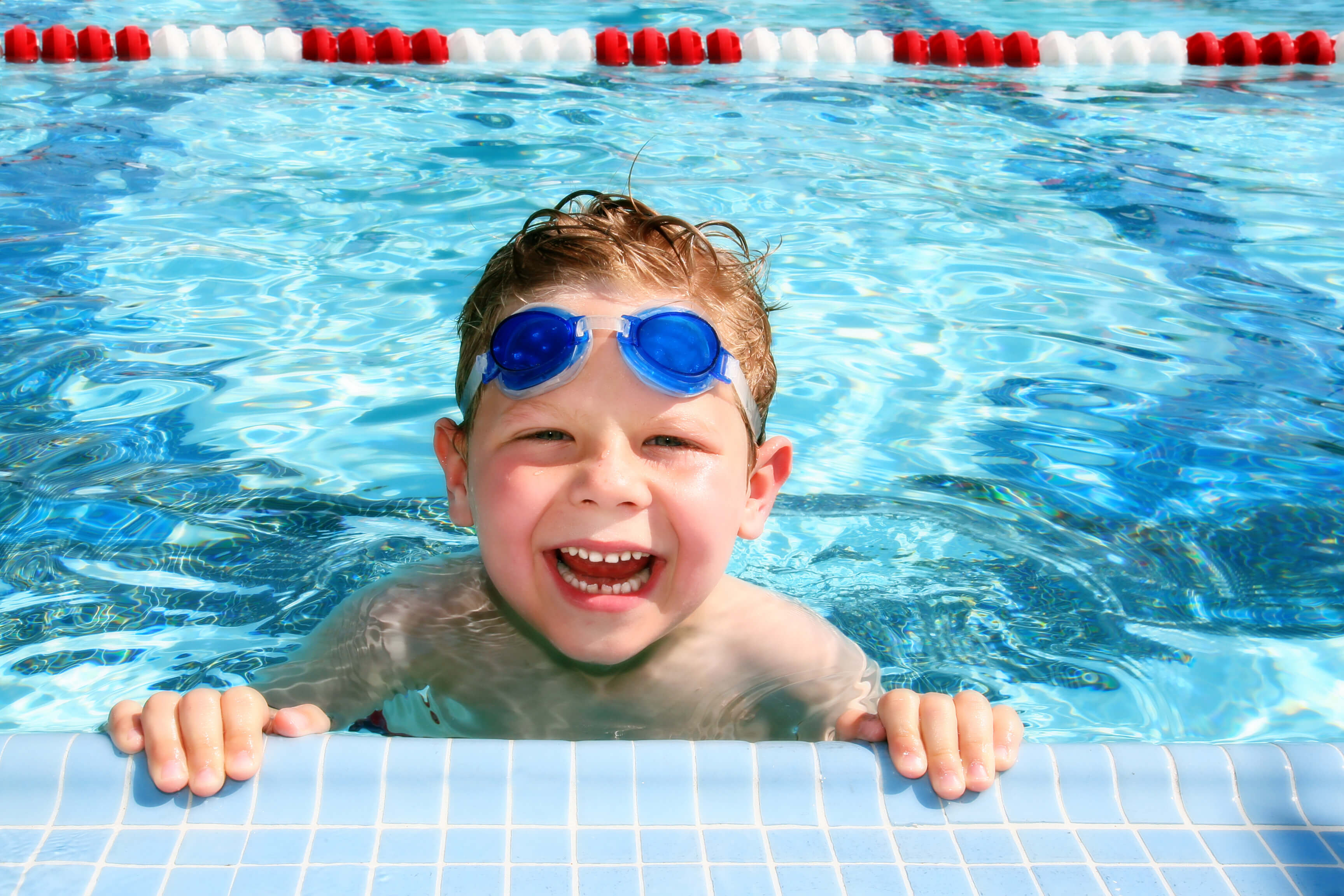 A young child with blue swim goggles smiles while holding onto the edge of a swimming pool in Huntington Beach.