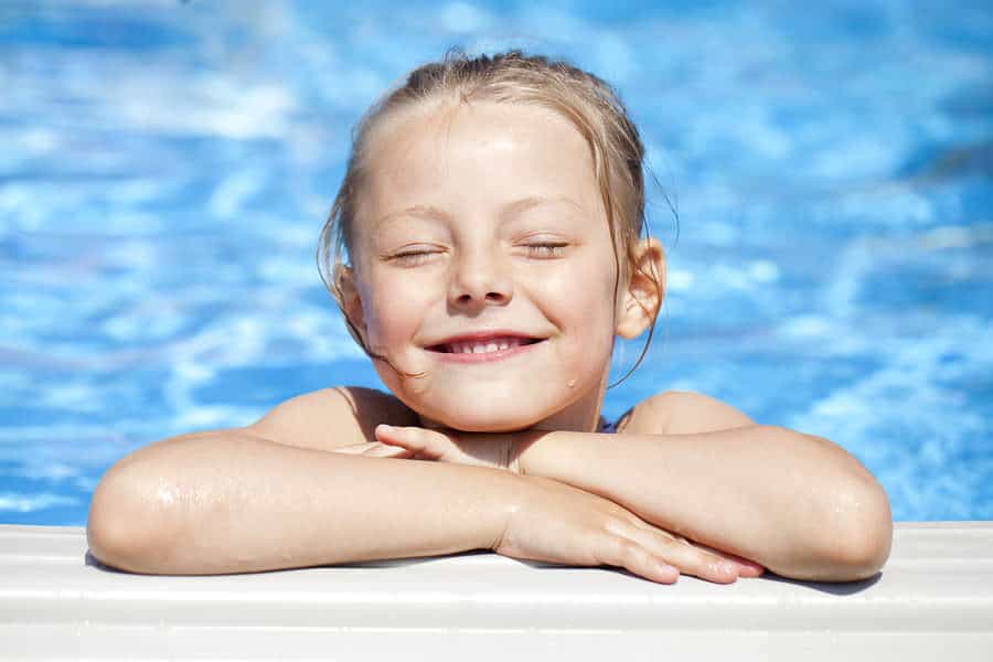 A young girl smiles with closed eyes, resting her chin on her arms at the edge of a swimming pool on a sunny day.