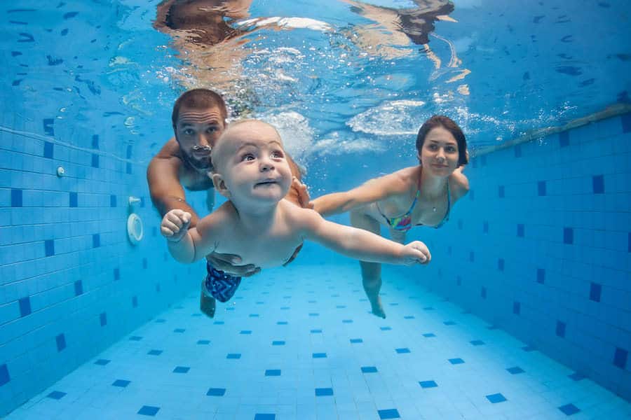 A young child swimming underwater in a pool, flanked by a man and a woman, all looking towards the camera.