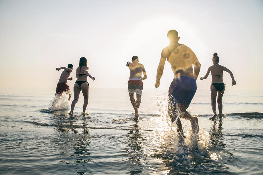 Group of people running into the sea at sunset, splashing water around, viewed from behind.