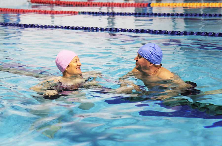 Two elderly people, a man and a woman wearing swim caps, happily talking in a swimming pool with lane ropes in the background.