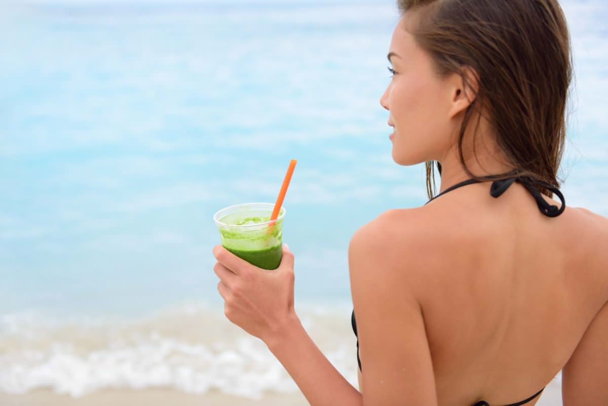 Woman in a black swimsuit holding a green nutrition tips smoothie on a beach, looking over her shoulder at the sea.