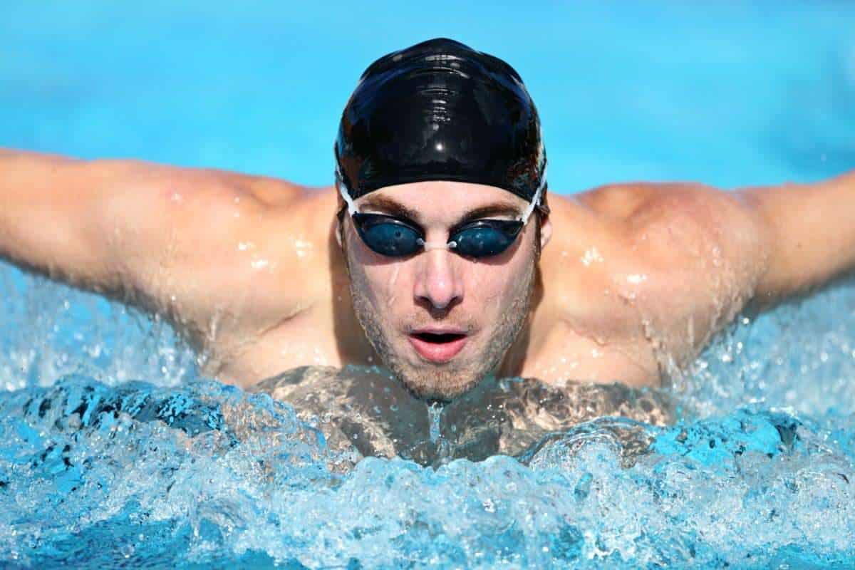 A male swimmer wearing goggles and a swim cap is swimming the butterfly stroke with bilateral breathing in a clear blue pool.