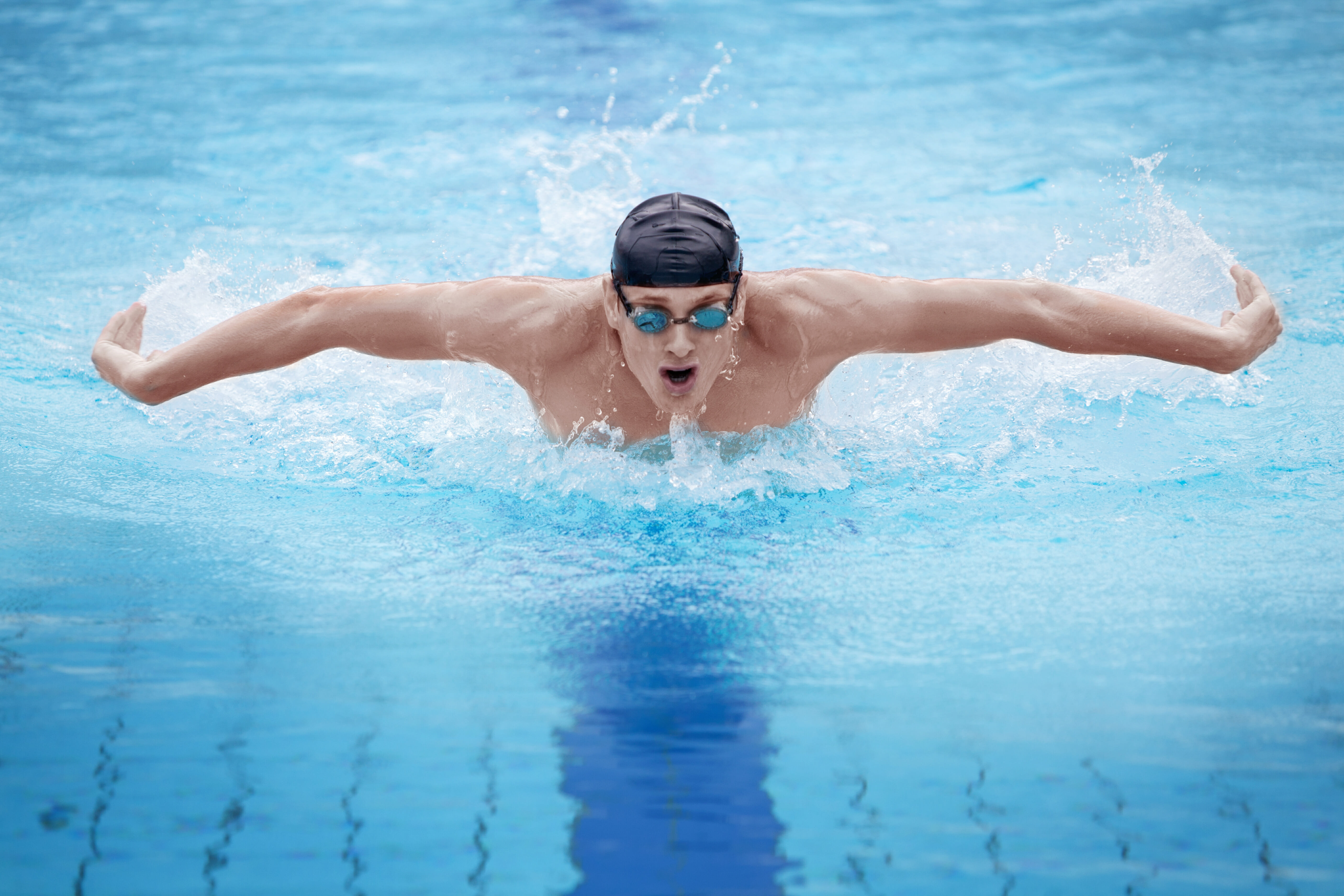 A male swimmer in goggles and a swim cap performing the butterfly stroke in a pool, creating splashes around him.