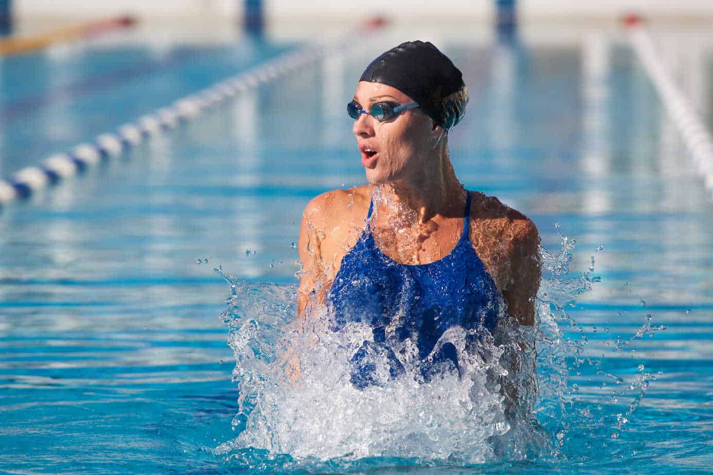 A female swimmer in a blue swimsuit and swim cap, emerging from the water in an outdoor pool during a swim.