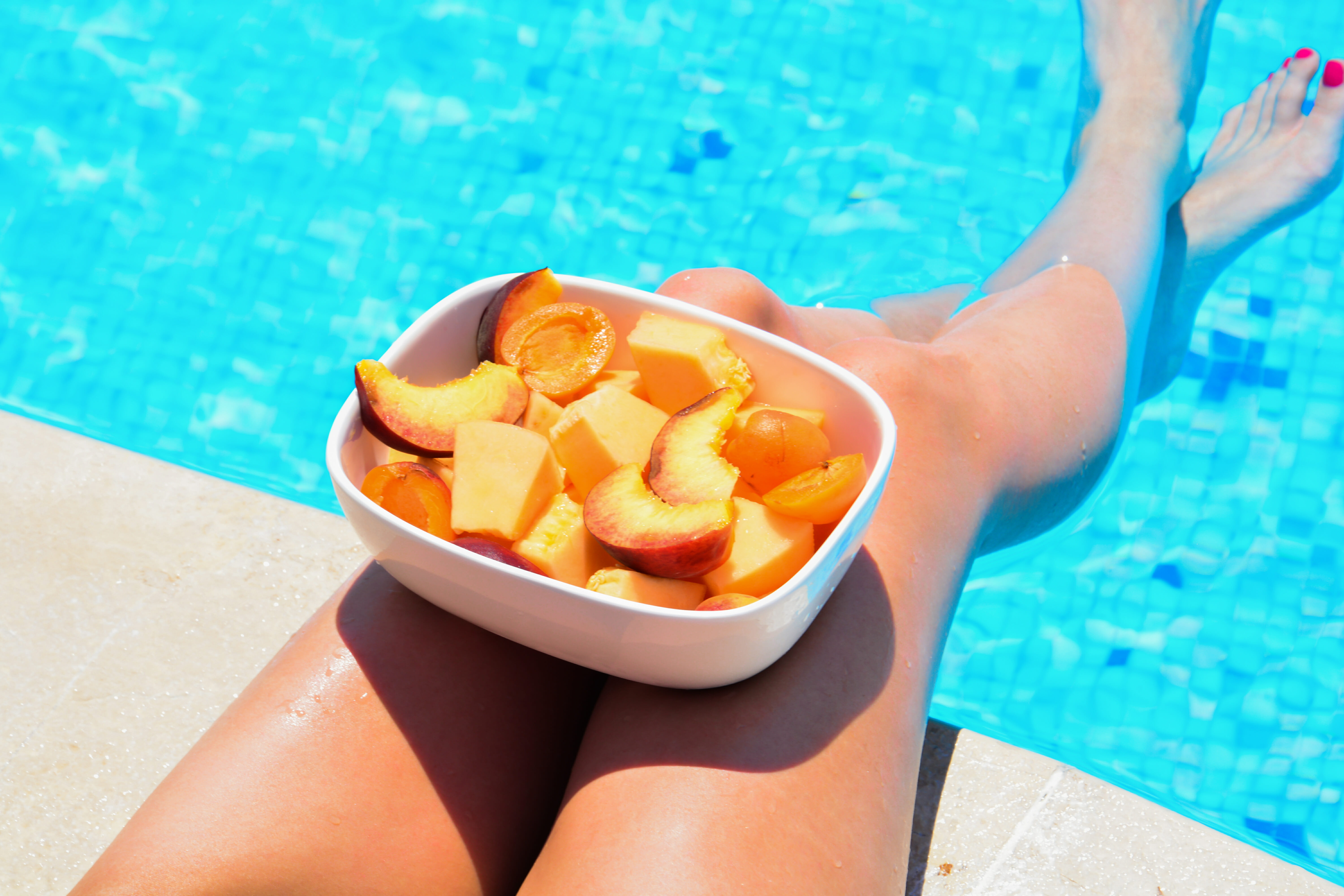 A person relaxing by a pool with a bowl of sliced peaches on their lap, showcasing a close-up of the legs and the colorful pool water.