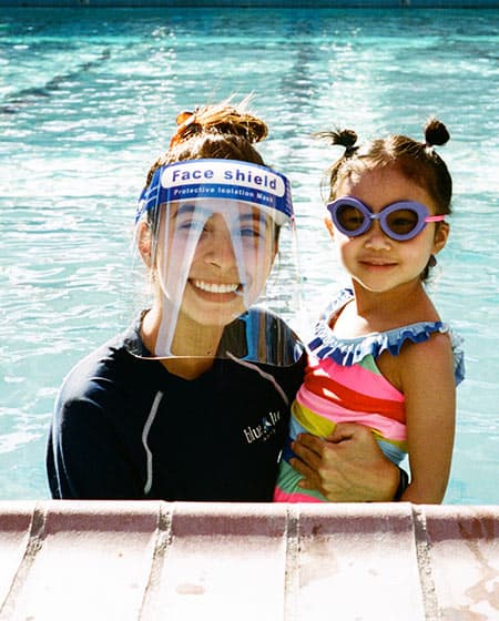A woman wearing a face shield and a young girl in purple goggles smiling together in a swimming pool.