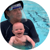 A man wearing a swim cap and goggles holds a laughing baby in a swimming pool.