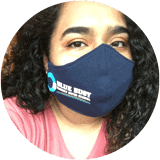 A woman with curly hair wearing a blue mask and pink top, with a logo and text on her mask.