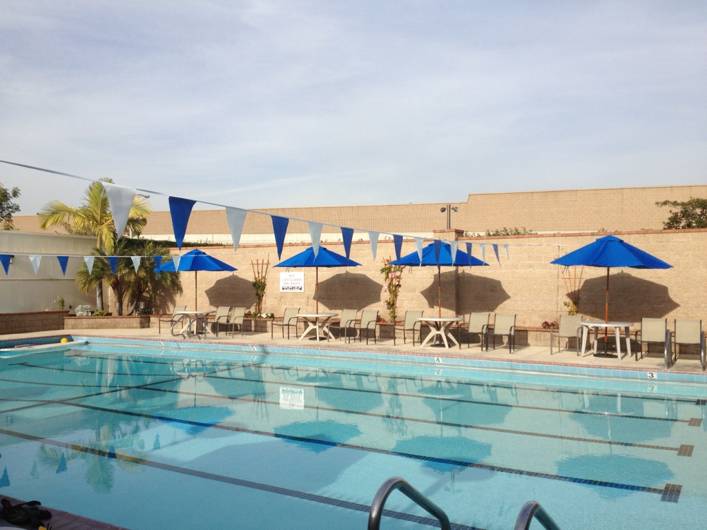 Outdoor swimming pool with blue umbrellas, lounge chairs, and triangular flags, reflecting a clear sky.