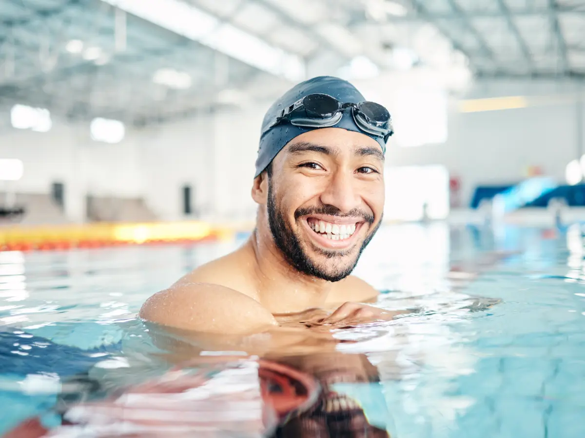 A swimmer with a beard and swim cap smiles in a pool, with the background showing a bright indoor swimming facility.