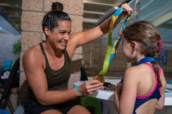 A woman shows a young girl a medal at a swimming pool, both smiling as the woman holds the medal up for the girl to see.