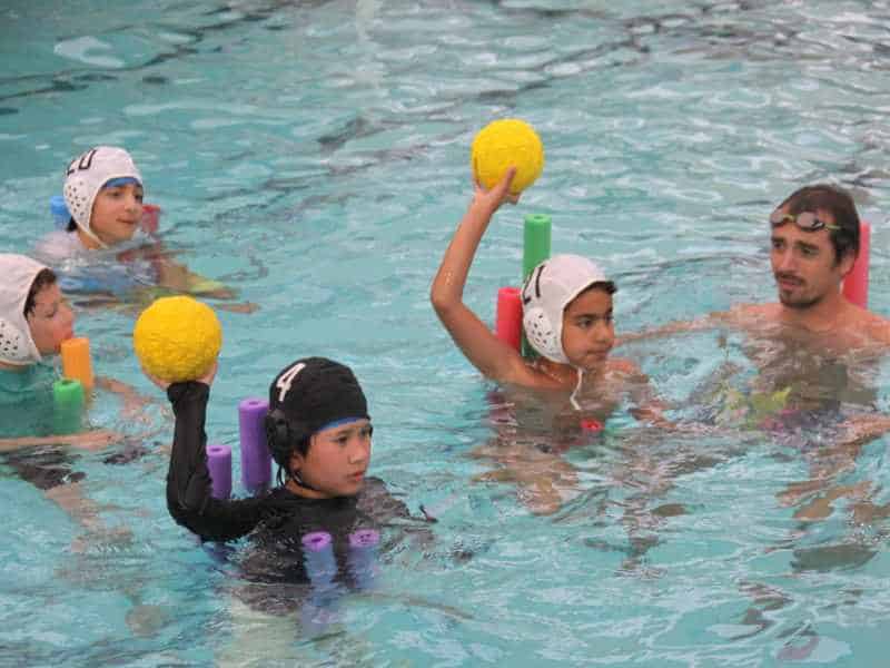 A group of children learning water polo techniques in a pool with an instructor.